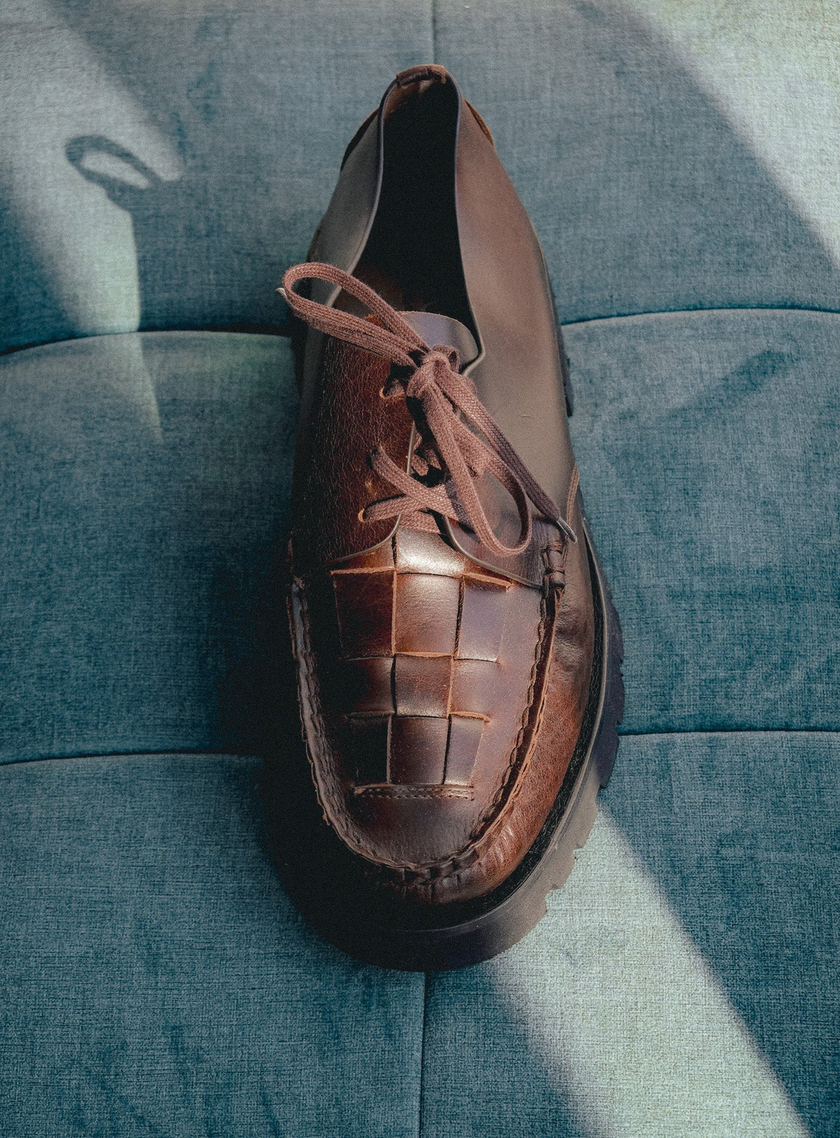 A brown leather lace-up shoe with a woven basket-like pattern on the toe, resting on a textured blue fabric surface with dramatic diagonal shadows.